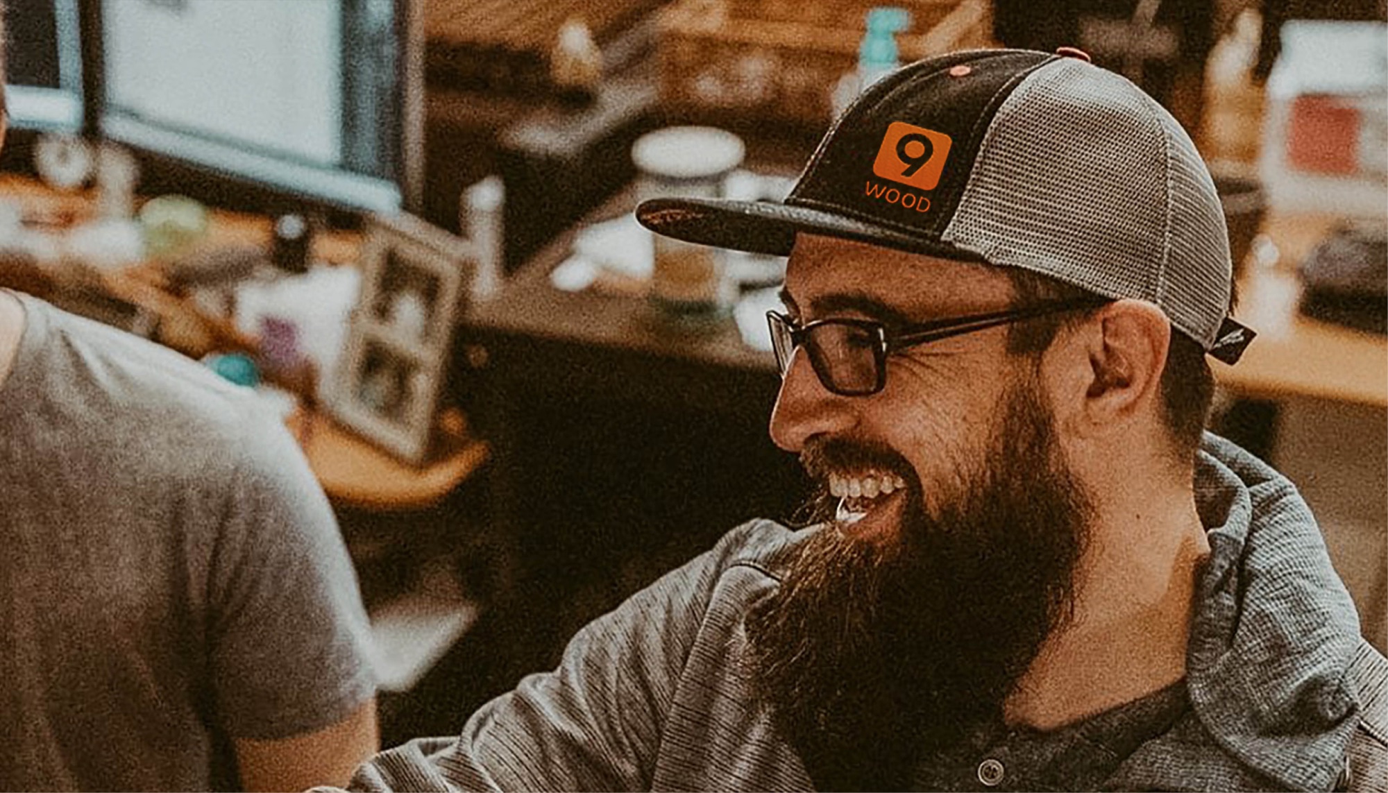 Workshop portrait: a bearded staff member in glasses and a 9Wood trucker cap laughing with a colleague on the shop floor