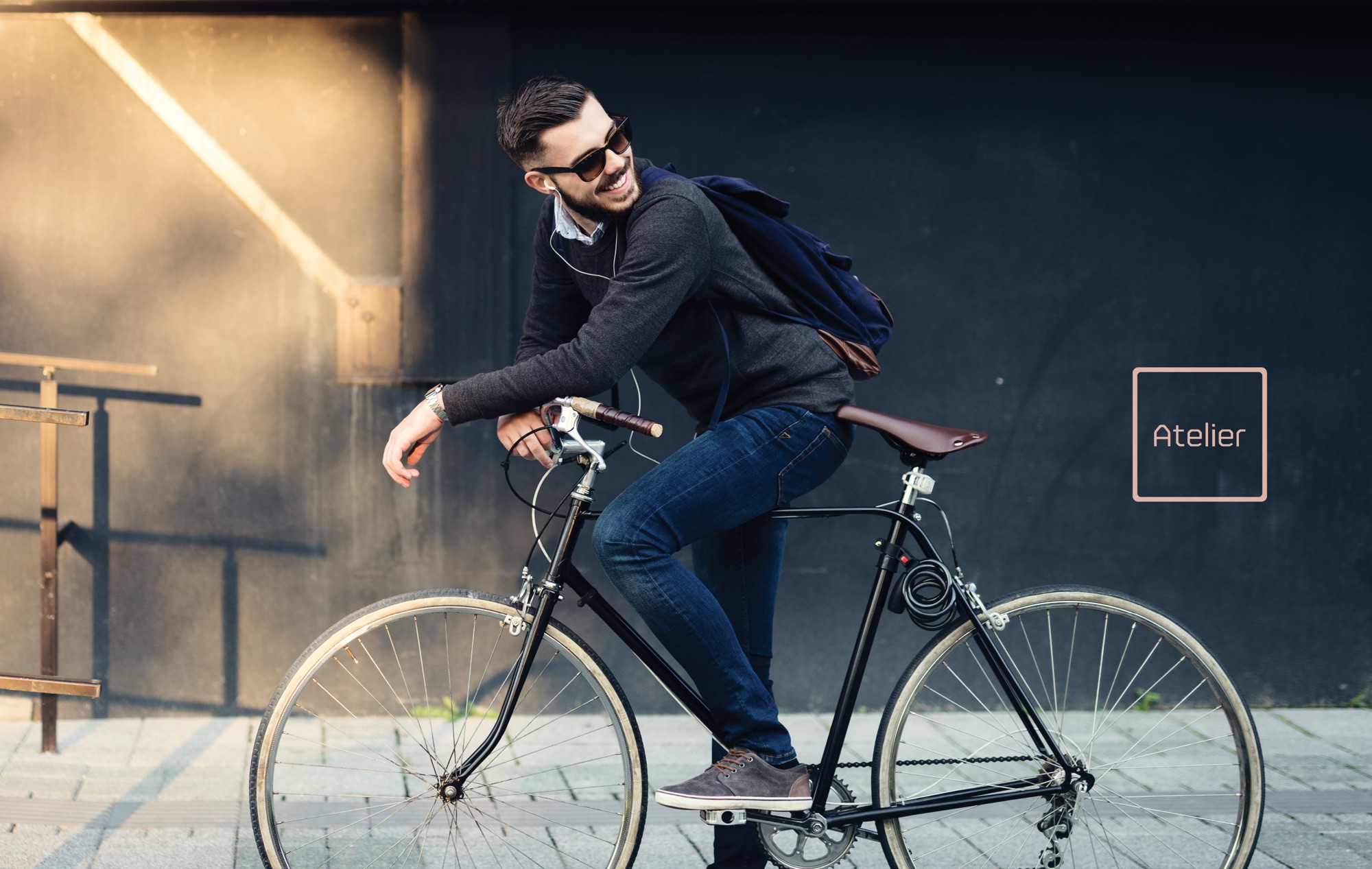 A man in sunglasses and a grey sweater pausing on a black fixed-gear bicycle against a dark wall in low sun, with the Atelier square logo in blush at the right