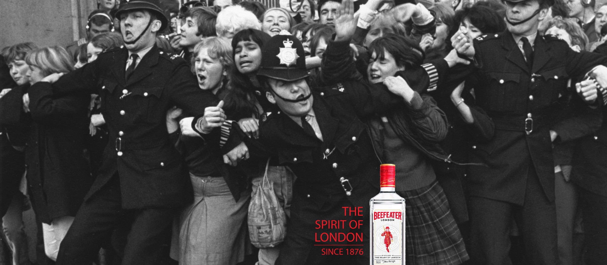 A black and white press photograph of police linking arms to hold back a crowd of screaming fans, a Beefeater bottle composited at the right beside “The Spirit of London, since 1876” in red