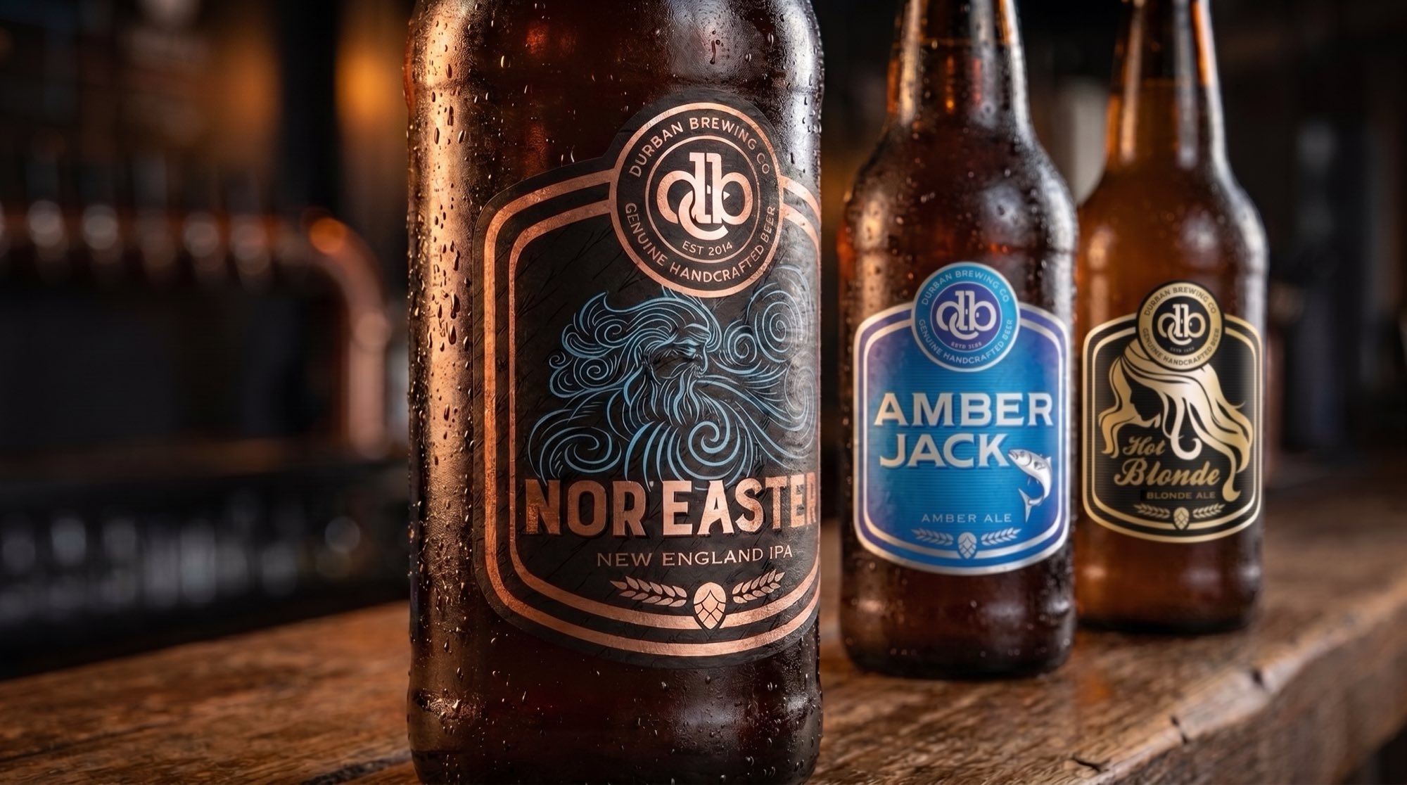 Three Durban Brewing Co. bottles on a bar counter — Nor’Easter New England IPA in front, its copper-on-black label carrying a blue line-drawn storm god, with Amber Jack amber ale in blue and Hot Blonde blonde ale in gold behind