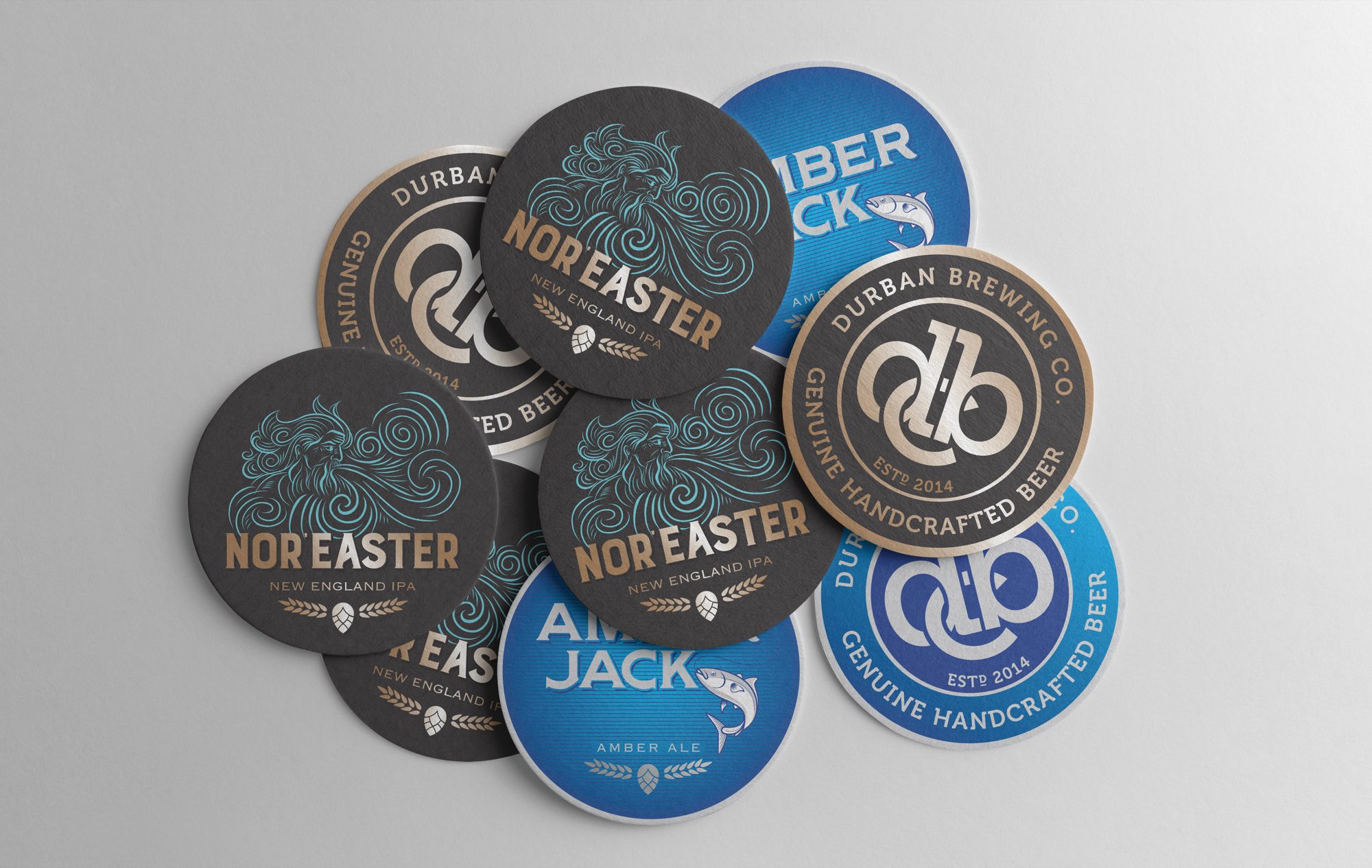 A scatter of Durban Brewing Co. beer mats — Nor’Easter in black and copper, Amber Jack in blue, and the dlb roundel reading Durban Brewing Co., Genuine Handcrafted Beer, Estd 2014