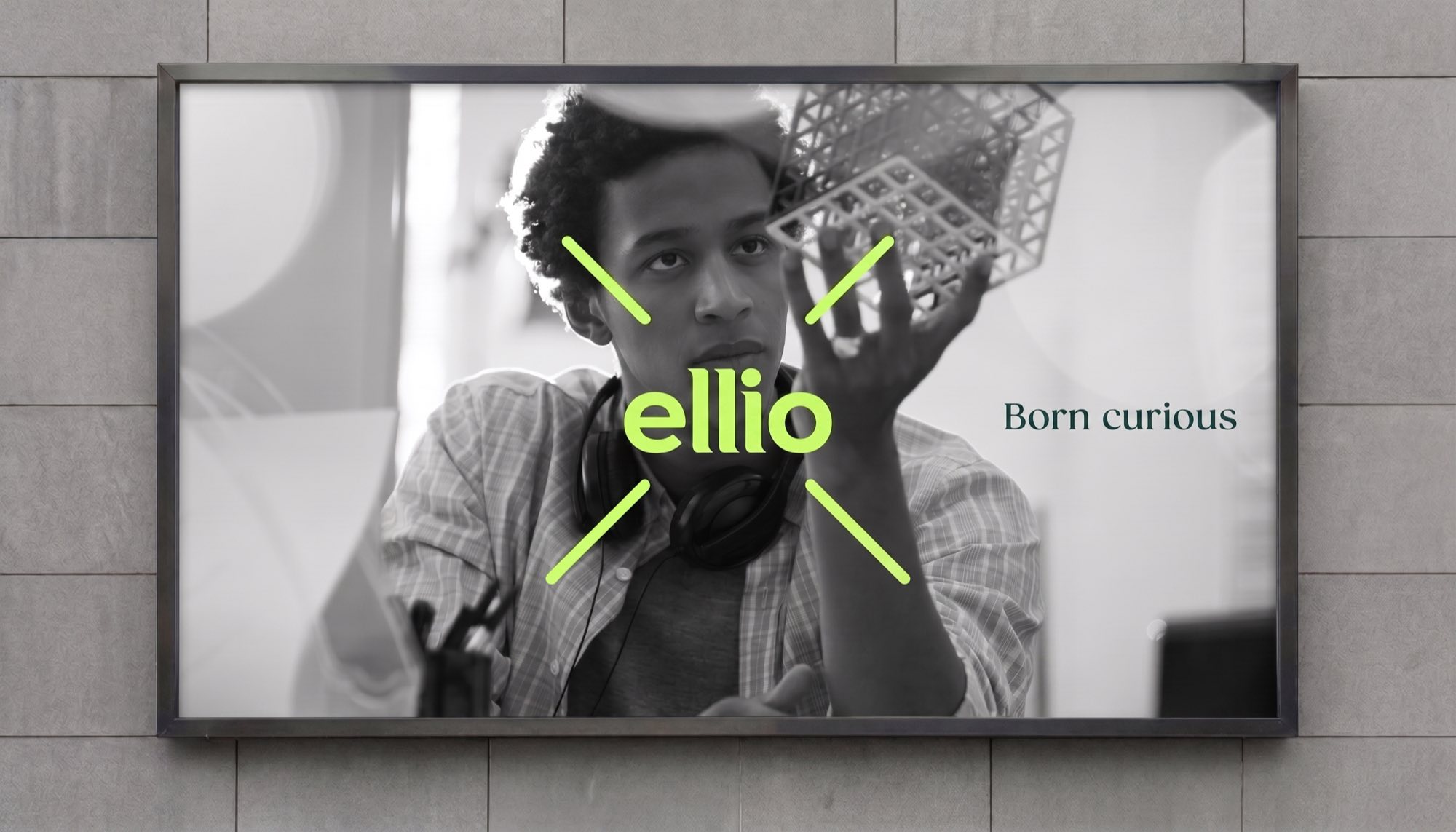 A backlit poster on a tiled wall: a black and white photograph of a young man examining a lattice structure, the ellio logo in acid lime at the centre of four angled strokes, with “Born curious” in a serif alongside