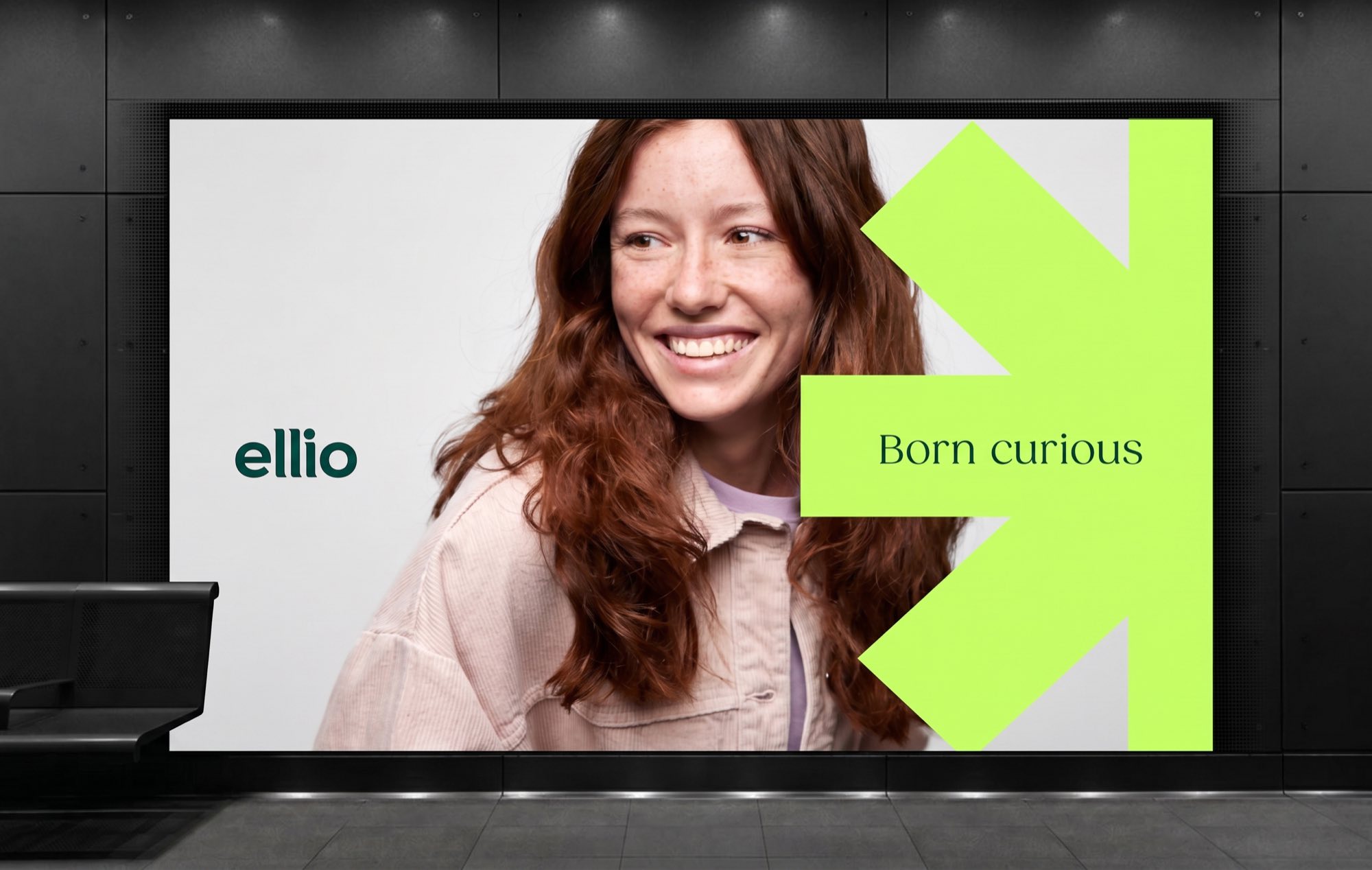 A station billboard: a laughing woman in a pink jacket, the ellio wordmark at the left, and “Born curious” set in a serif on an acid lime arrow device