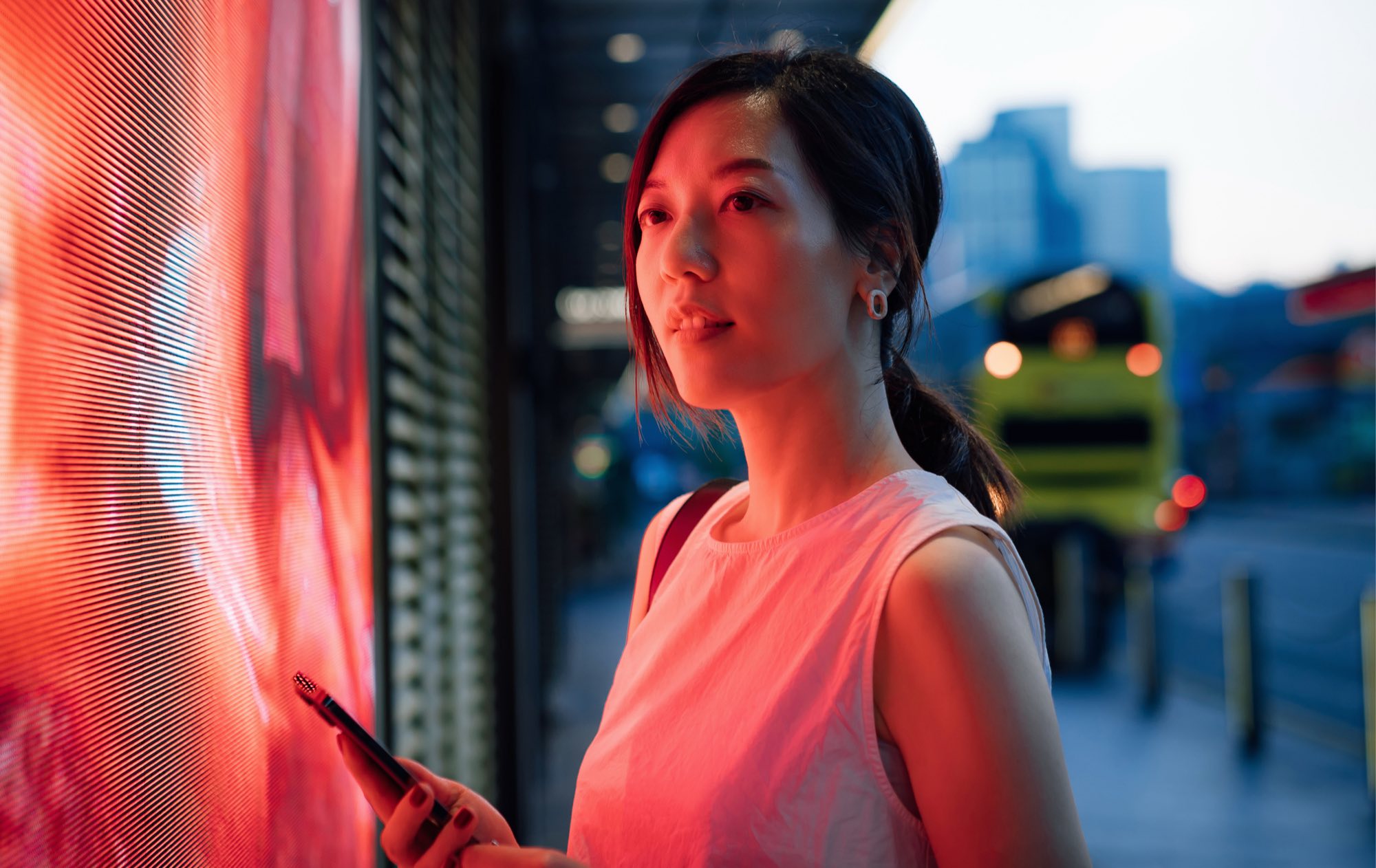 A woman at a large LED screen at dusk, lit red by the display, phone in hand, with a bus and city buildings behind