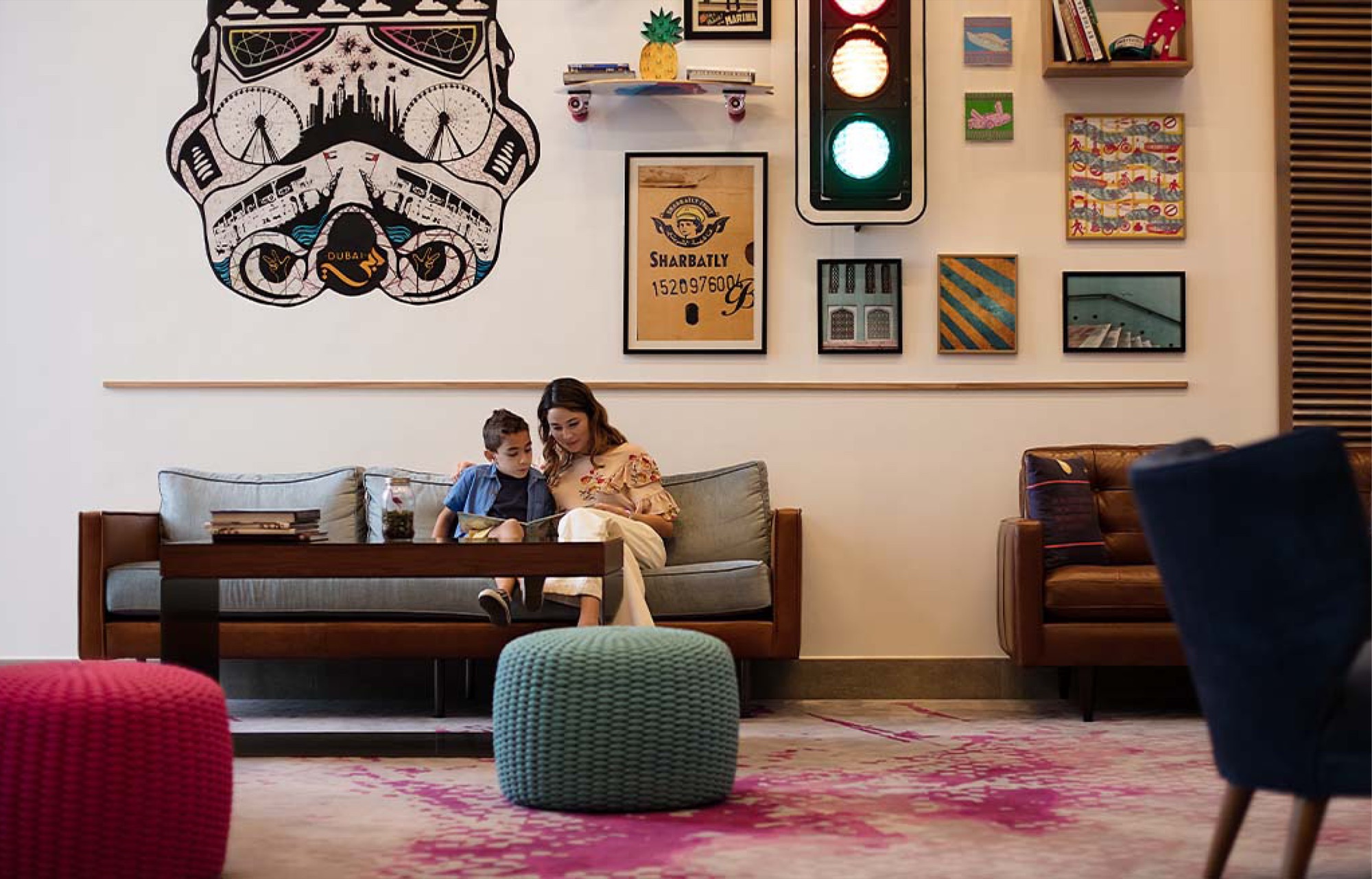 Lobby lounge: a mother and child reading on a leather and linen sofa beneath a large stormtrooper mural drawn from Dubai landmarks, flanked by framed prints, a vintage traffic light and a skateboard shelf, with knitted pink and teal poufs on a magenta-splashed rug