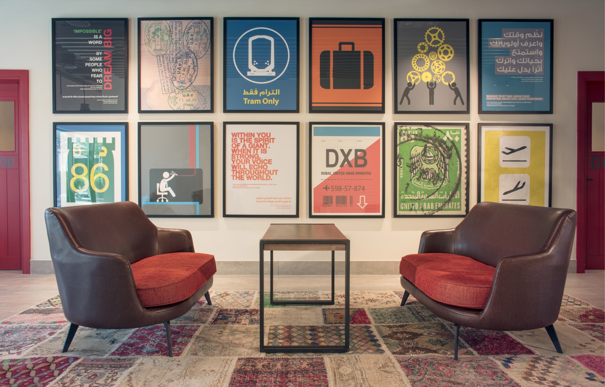 Poster wall between two red doors: twelve framed graphics including a DXB baggage tag, a UAE passport stamp, a Tram Only sign, a suitcase icon, and quotation posters in English and Arabic, above two brown leather armchairs with red cushions on a patchwork kilim