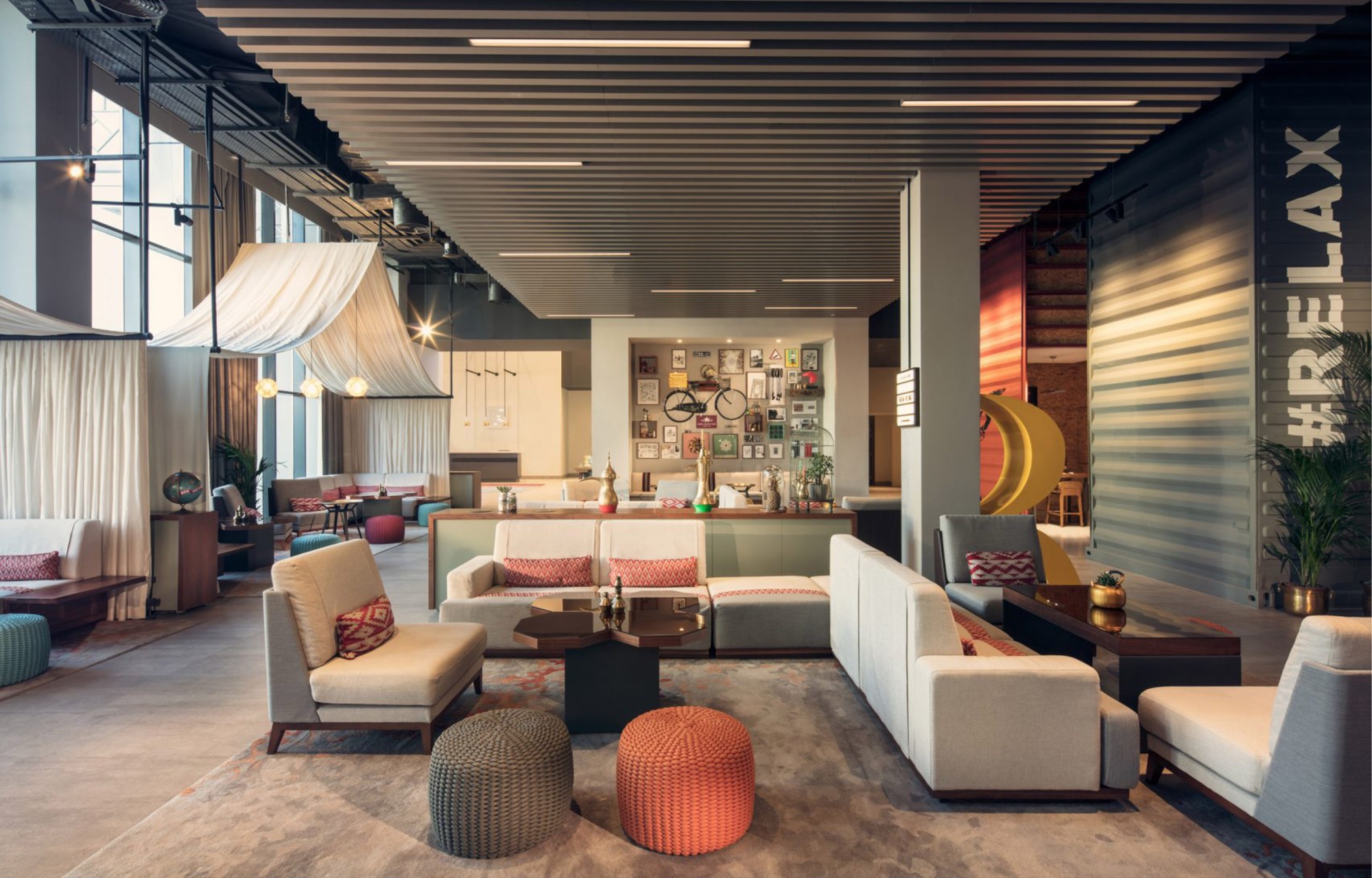 The Daily restaurant: pale timber tables and sage metal chairs under oversized yellow woven pendants, with a wall of scattered patterned ceramic plates, a glazed convenience store beyond and a fretwork screen to one side