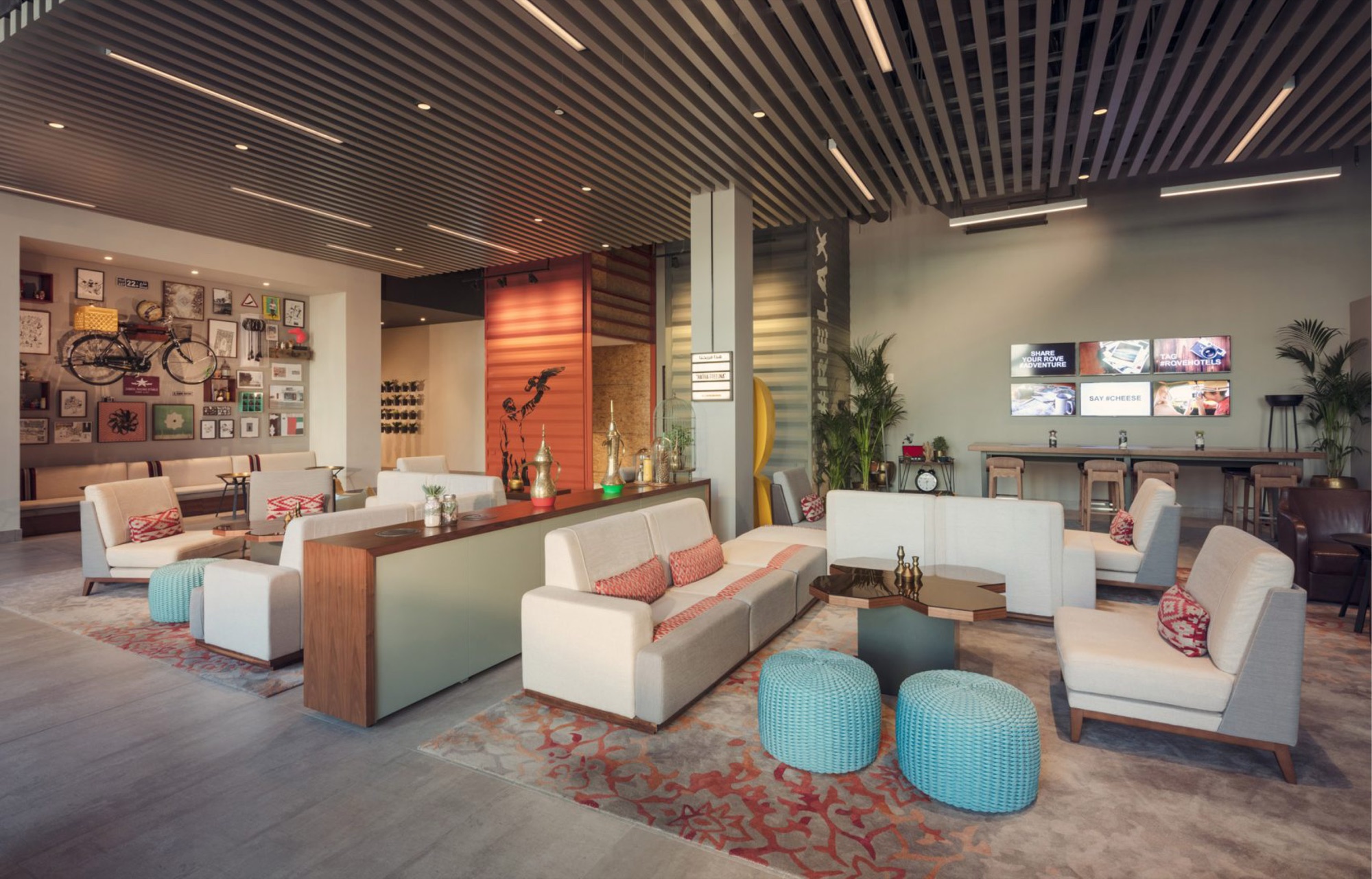 Social lounge: low cream seating with red ikat cushions around a brass-topped octagonal table, a long communal bar bench under a bank of screens reading Share your Rove adventure and Tag #ROVEHOTELS, with the bicycle gallery wall and orange container facade behind
