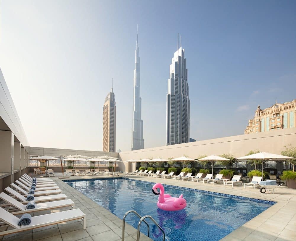 Rooftop pool in daylight: rows of white loungers with rolled towels and parasols along a rectangular pool holding a single pink flamingo float, the Burj Khalifa and neighbouring towers rising behind