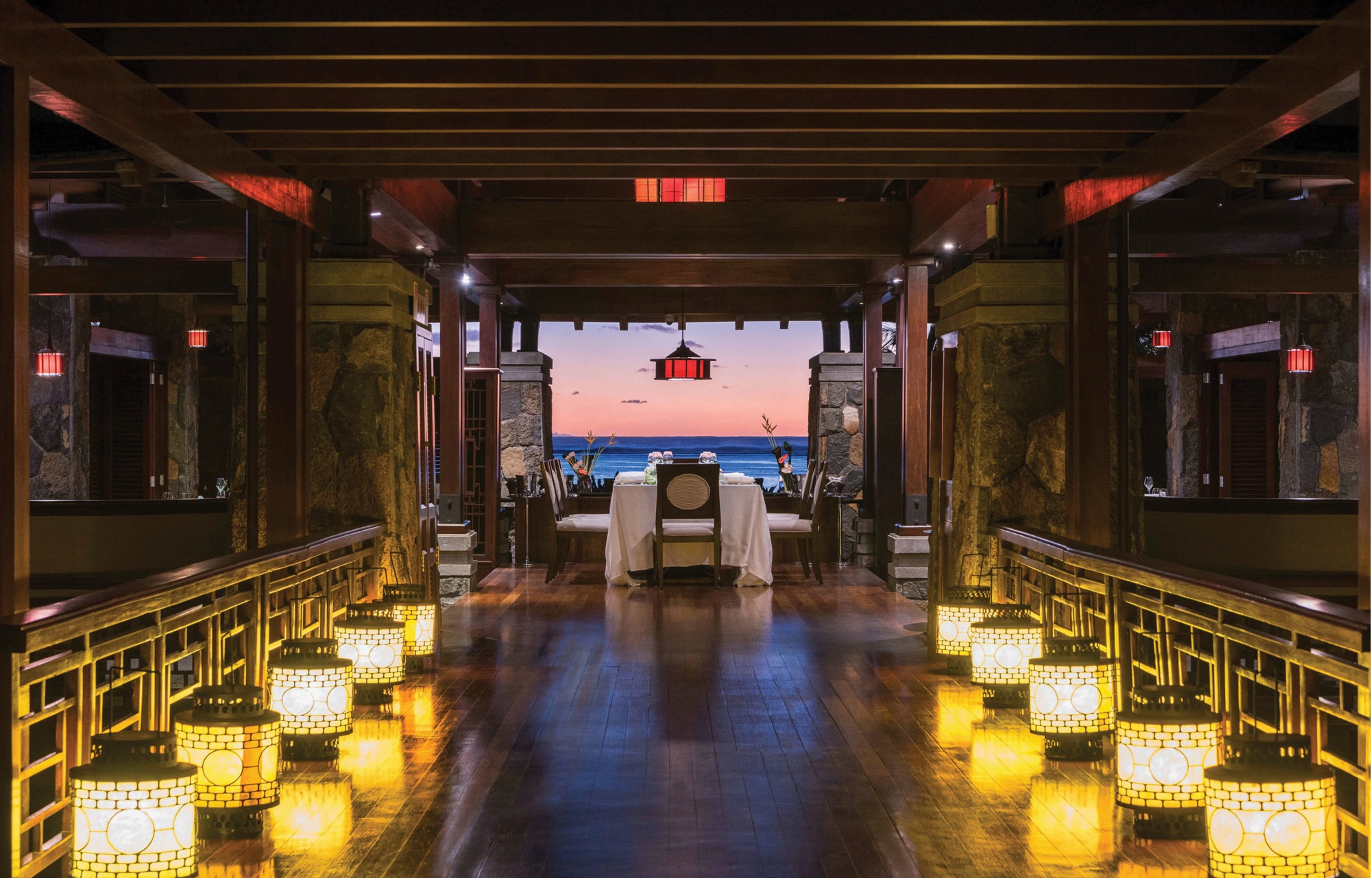 The Asian restaurant at dusk: a dark timber walkway lined with glowing stone lanterns leading to a laid table framed by the open pavilion, the ocean and a pink sunset beyond