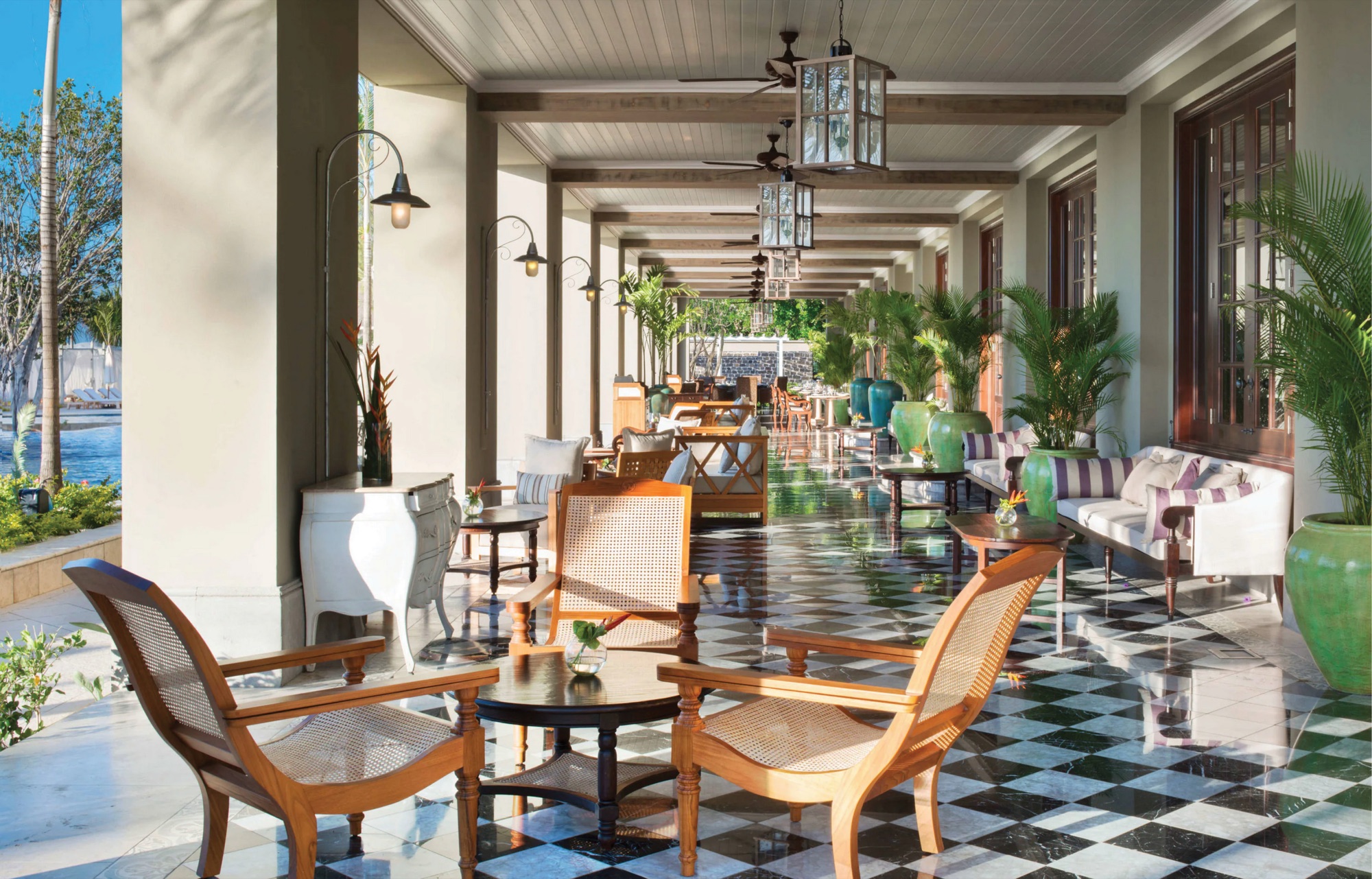 The manor house veranda: black and white chequered marble, caned planter chairs, white sofas with striped cushions, glazed green urns of palms, and lantern pendants running the length of the colonnade