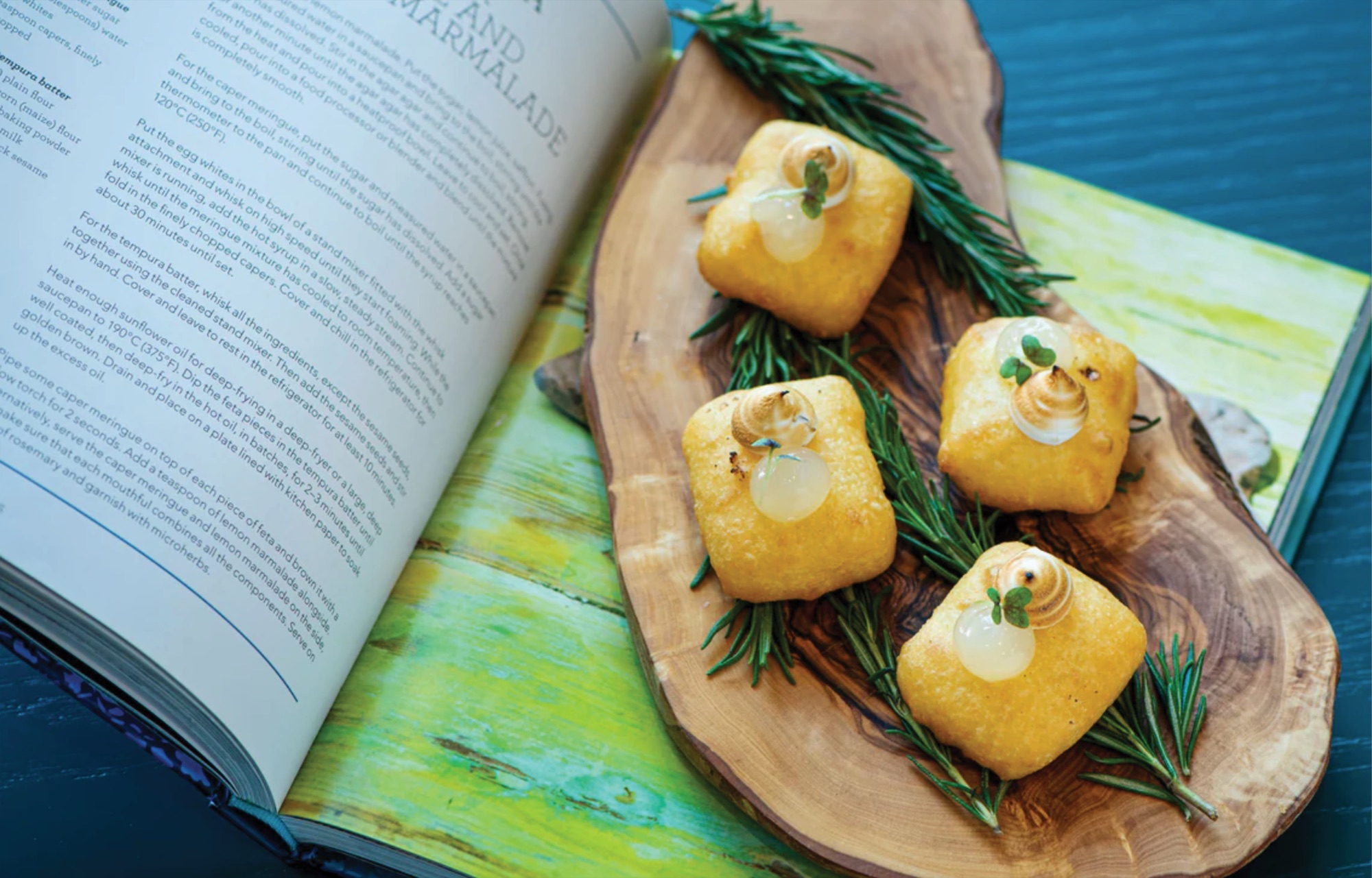 An open recipe book beside a live-edge olive board holding four squares of tempura feta, each topped with a torched caper meringue and a sprig of microherb on rosemary