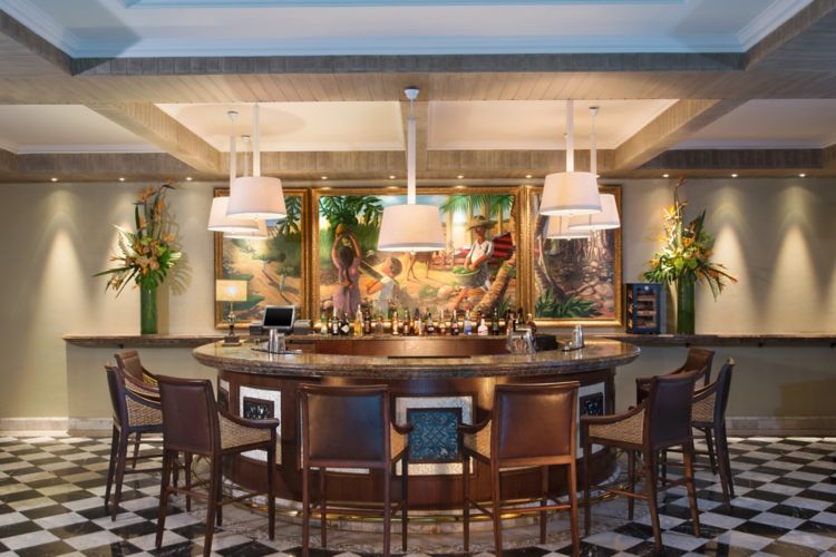 The bar: a circular timber and marble counter ringed with leather stools on a black and white chequered floor, drum pendants above, and a triptych of tropical paintings in gilt frames behind
