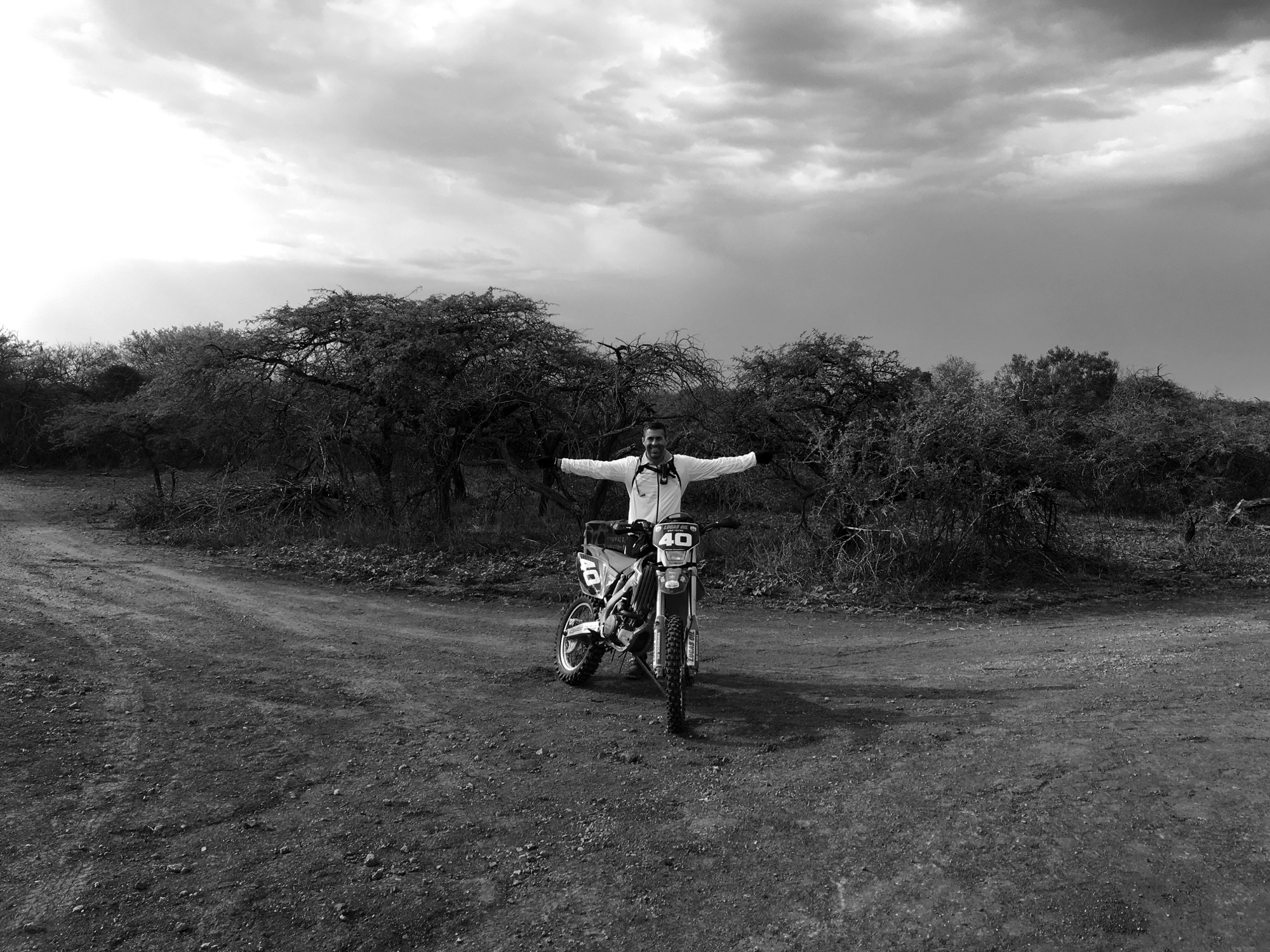 Paul Vosloo standing arms outstretched behind a number 40 enduro bike at a fork in a dirt road, thorn trees and a heavy cloudbank behind, in black and white