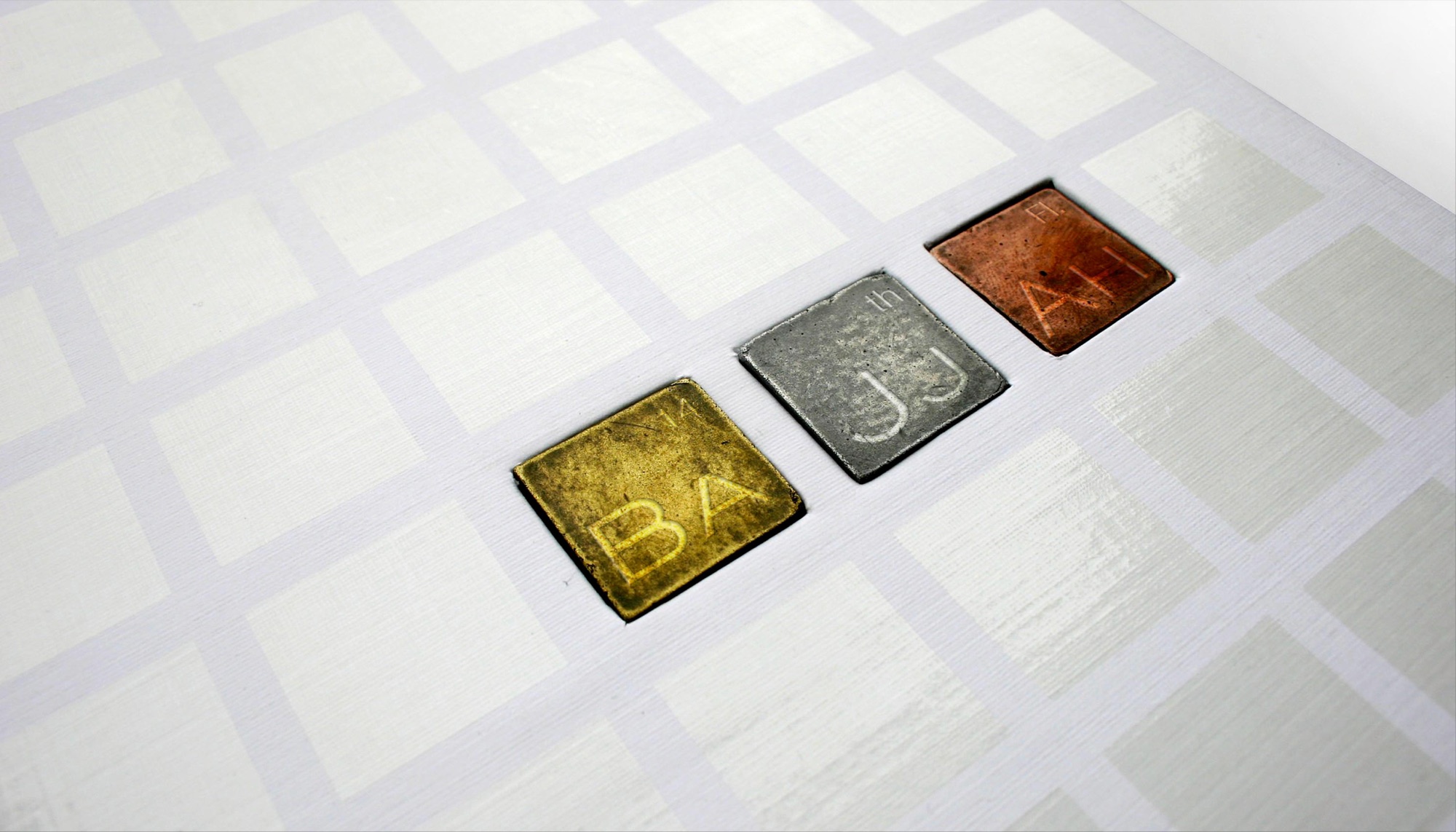 Room and floor signage detail: three small cast metal tiles — brass, pewter and copper — set flush into a white tiled surface, each engraved with an Arabic and Latin character pair