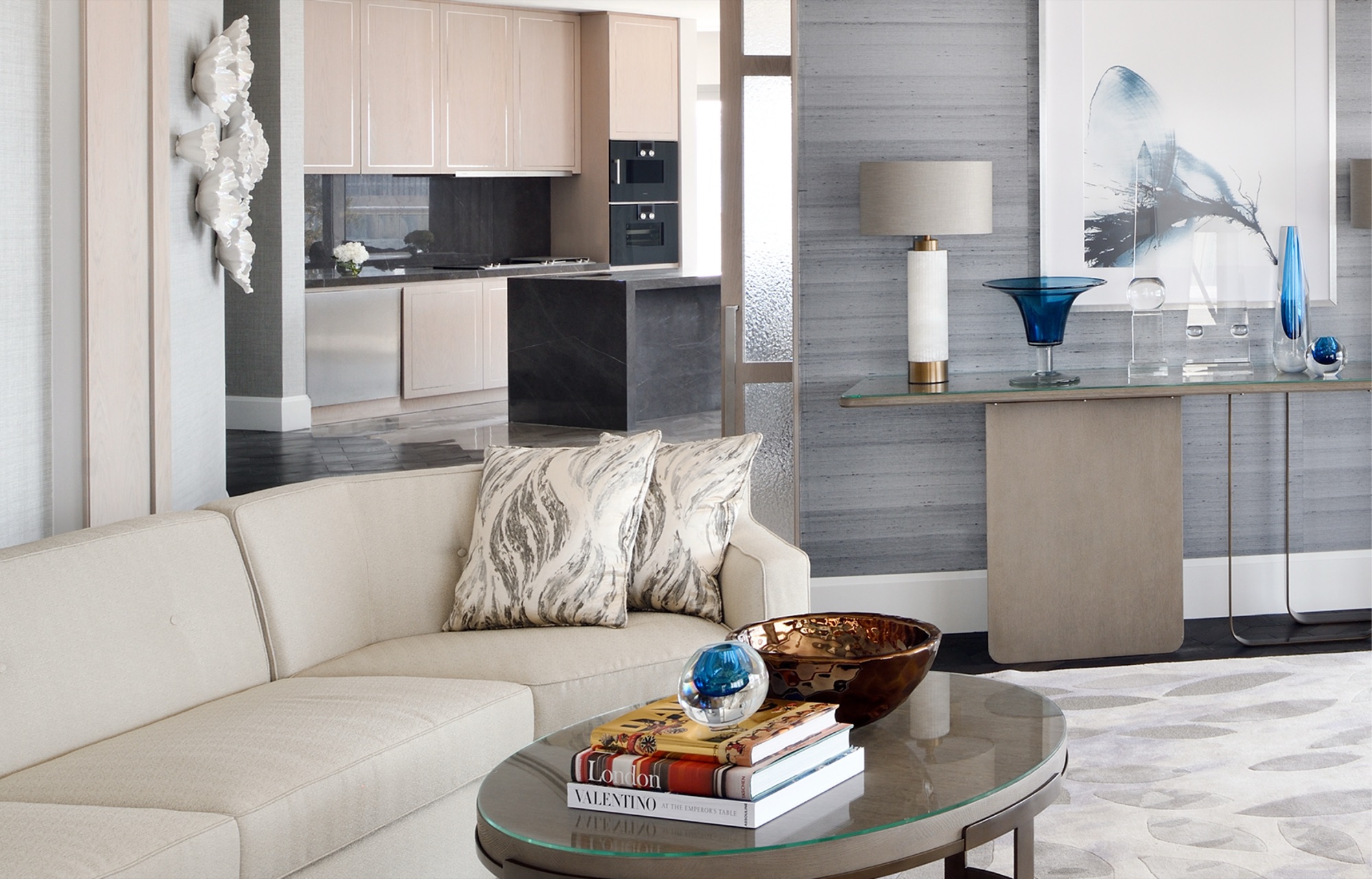 Serviced apartment interior: a curved cream sofa and oval glass coffee table, an open kitchen in pale oak with a dark stone island, a console of blue glassware beneath a framed ink artwork, and a white ceramic wall sculpture