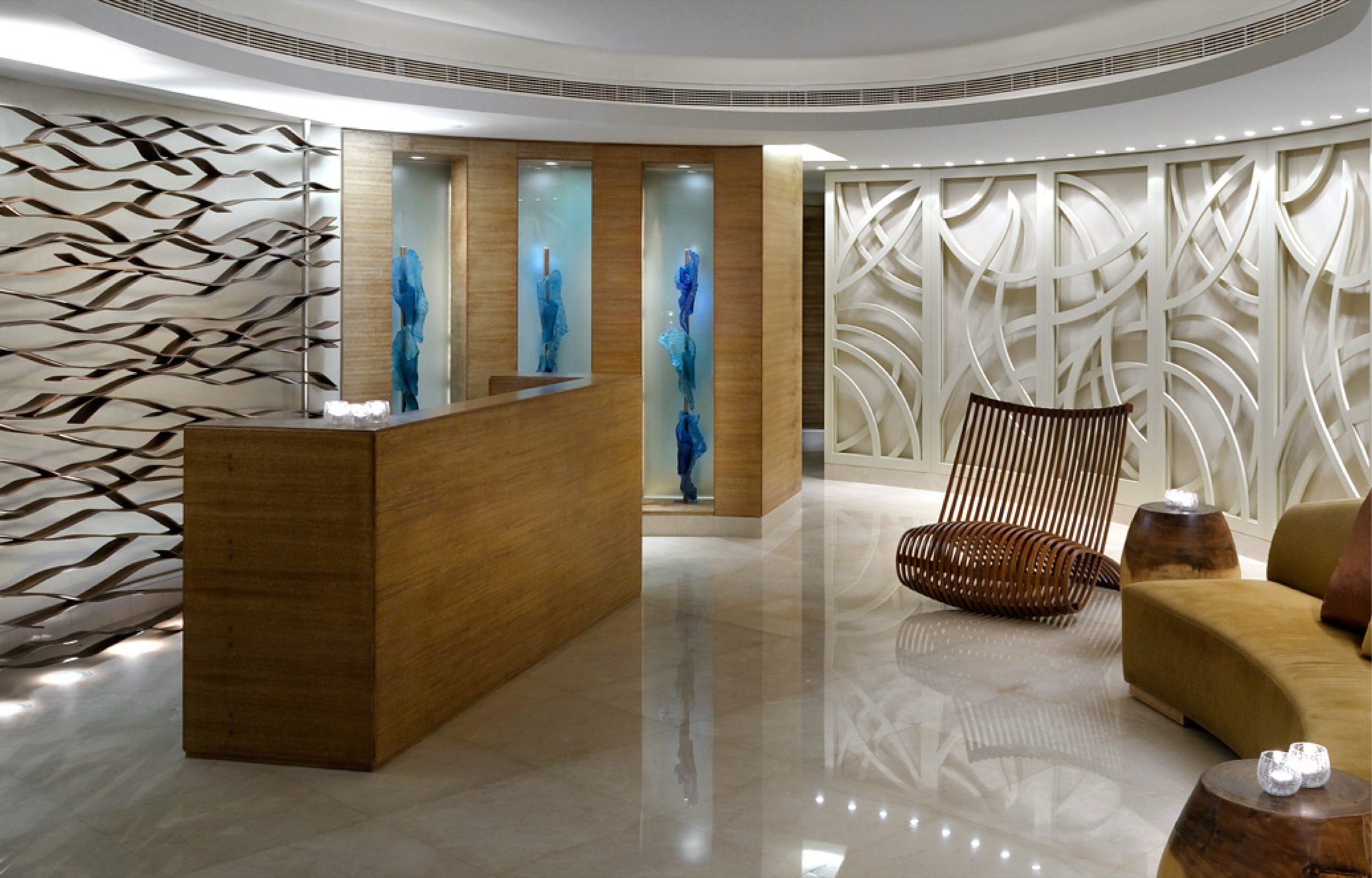Spa reception: a timber desk against a wall of layered bronze ribbons, three lit alcoves holding blue fabric sculptures, a curved white fretwork screen, a steam-bent timber lounge chair and candle-topped burl stools on polished marble